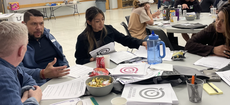 Workers sit at a table in a lunch discussion. There are "Secrets of a Successful Organizer" handouts with the bulleye logo on the cover, interspersed between a bowl of food, drinks and snacks. 