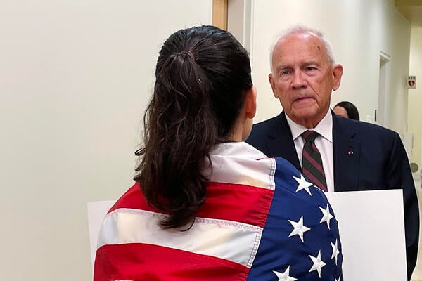 A photo of Robert Pence and Tehama Lopez in conversation. She has her back to the camera and an American flag wrapped around her shoulders.