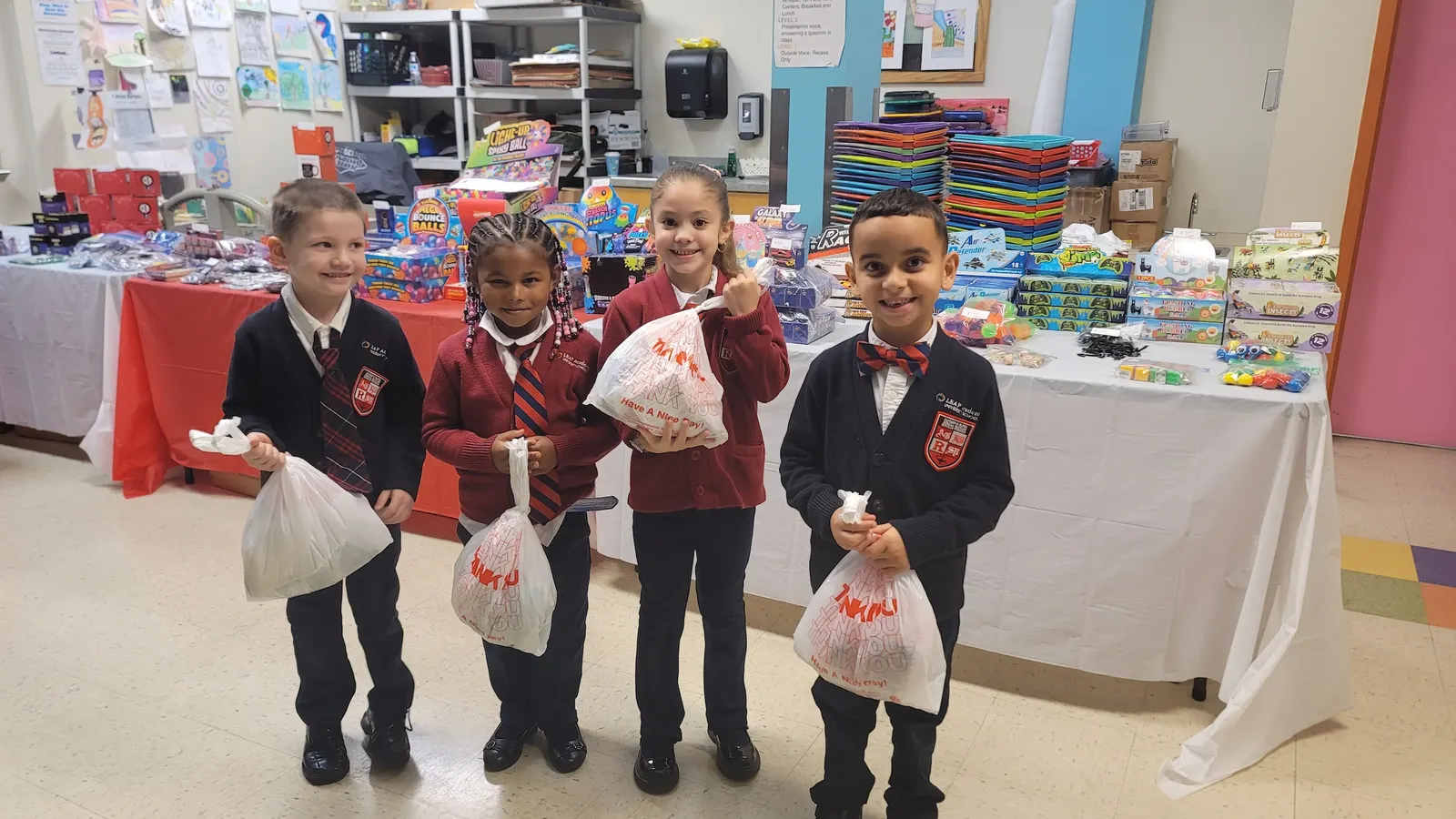 Four young students in school uniforms are holding plastic bags and looking at the camera. They are standing in front of a table with items.