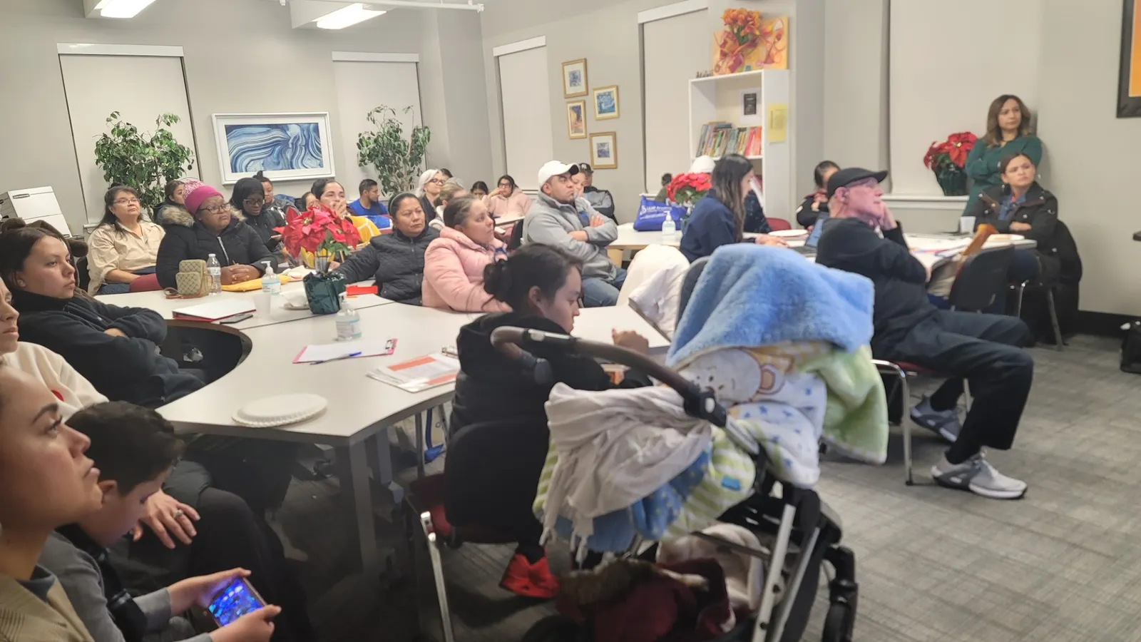 A room full of adults and a few children are sitting on chairs and at tables looking at a speaker.
