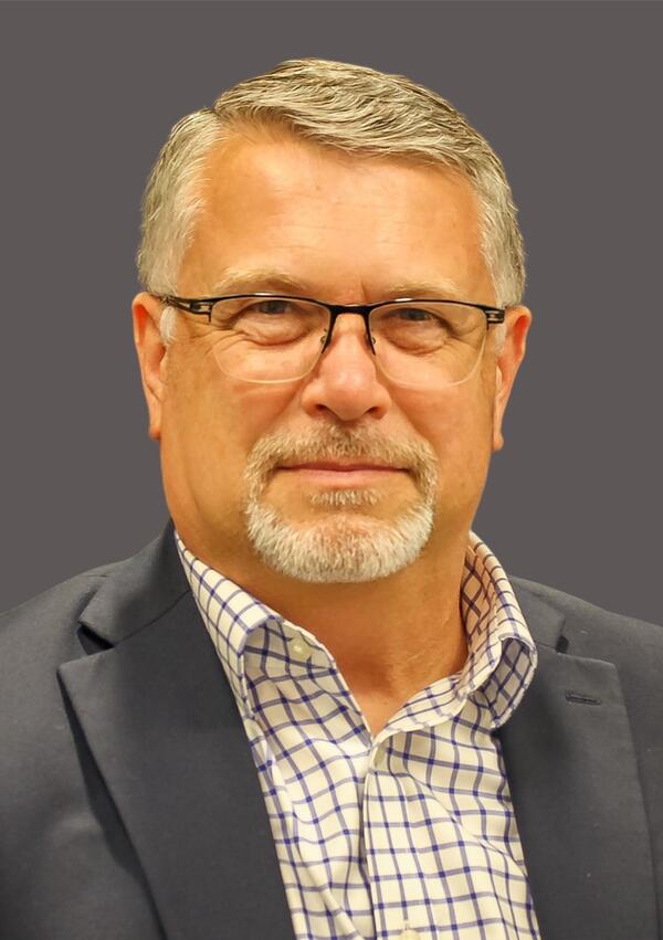 Dallas Kratzer poses for a headshot wearing a gray suit coat and checked collared shirt and glasses.