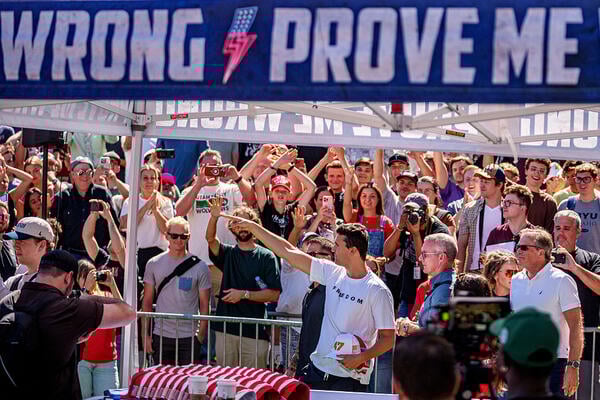 Charlie Kirk, in a white shirt, points to the crowd while holding some hats in his hand