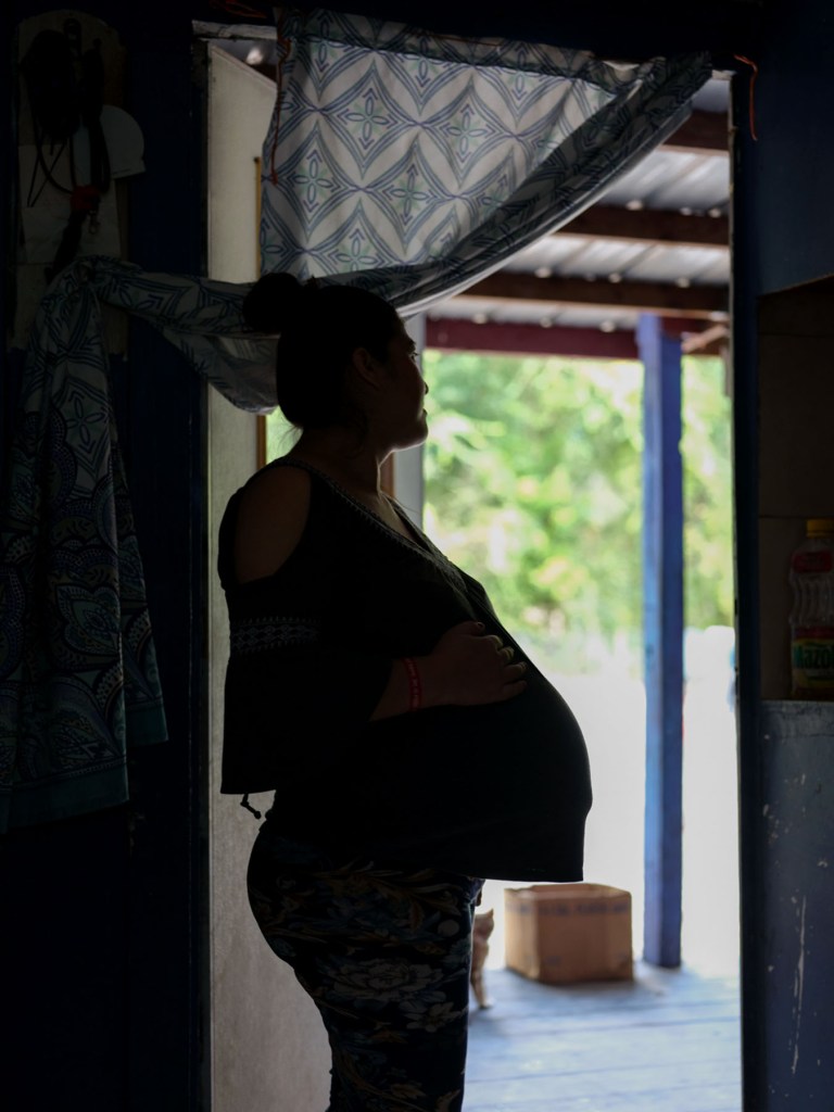 A pregnant woman stands in silhouette inside a dark doorway, holding her belly and looking outside toward the sunlight and trees.