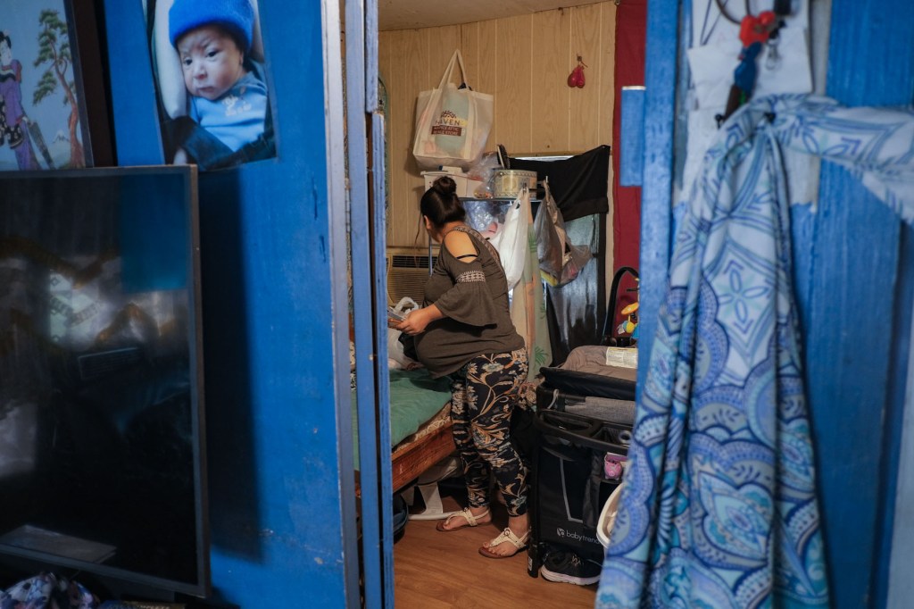A pregnant woman bends over a bed, sorting through baby items in a small, crowded bedroom with blue-painted door frames.