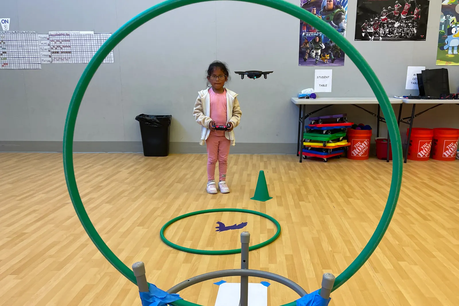 A student is holding a device while standing on a sports court inside. In the foreground is a hoop framing the photo from the camera.