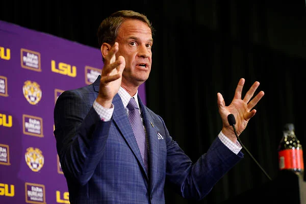 Lane Kiffin speaks at a press conference as he is introduced as the new head football coach of the LSU Tigers. He is a white man with short brown hair, wearing a blue suit with a purple tie and patterned shirt.