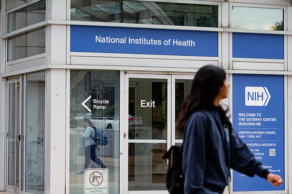 A pedestrian walks by a glass facade that says “National Institutes of Health."