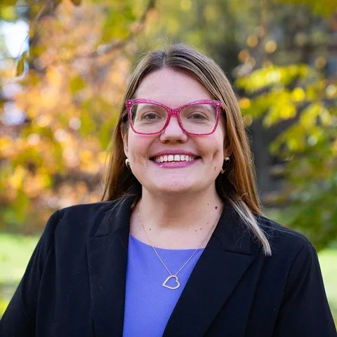 Kate Edwards smiles for a photo while wearing glasses, a heart necklace and a blazer.
