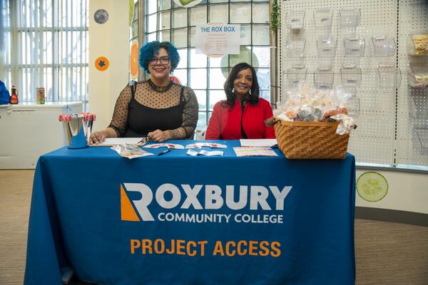 Two women, one with blue hair wearing a black dress with a sheer overlay, and one with shoulder-length dark hair wearing a red top, sit behind a table with a blue tablecloth that says "Roxbury Community College Project Access."