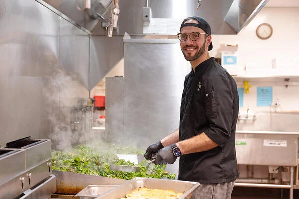 A light-skinned man in a black chef's coat, backward baseball cap and black gloves stands in front of a large flattop cooking surface stirring a pile of greens.