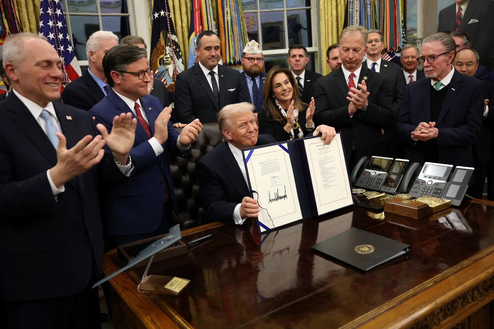President Donald Trump sits in the center of a group of people standing up and clapping around him as he holds up a folder with signed legislation.