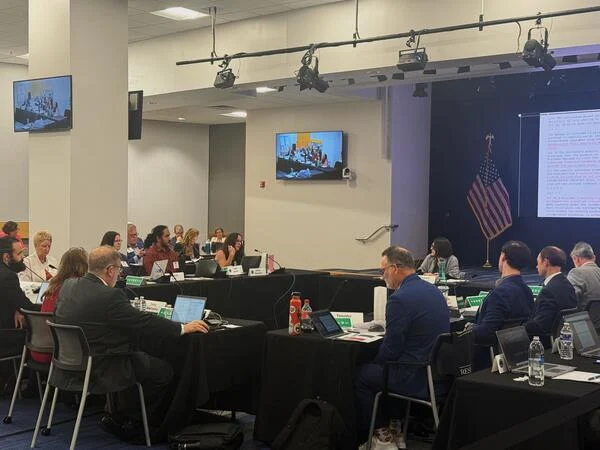 Committee members seated at four rectangular tables arranged in a square, covered with black tablecloths. Most have laptops in front of them.